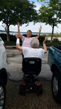 Traveling with Parkinson's | Parkinson's News Today | Bev, seated in her mobility scooter, is stopped between two cars in a parking lot. She's facing her sister, Bev, and both women have their arms raised in celebration while on vacation in Colonial Williamsburg, Virginia.