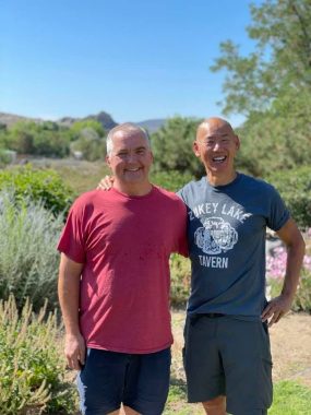 Parkinson's News Today | Brian Reedy, in a red T-shirt and shorts, poses with Jimmy Choi, in a blue-gray T-shirt and shorts, outdoors among a prairie field