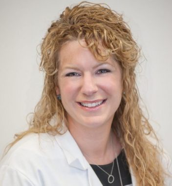 A headshot image of a woman with long blonde hair. She wears a white coat with a black shirt beneath, as well as a necklace. 