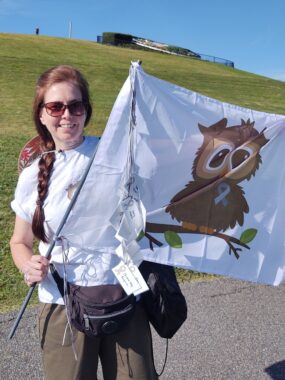 A woman with reddish brown hair in a long pigtail stands on a road with a hill of green grass behind her. She wears a white T-shirt and brown paints and carries a mostly white flag, featuring the image of a cartoon owl on a branch.