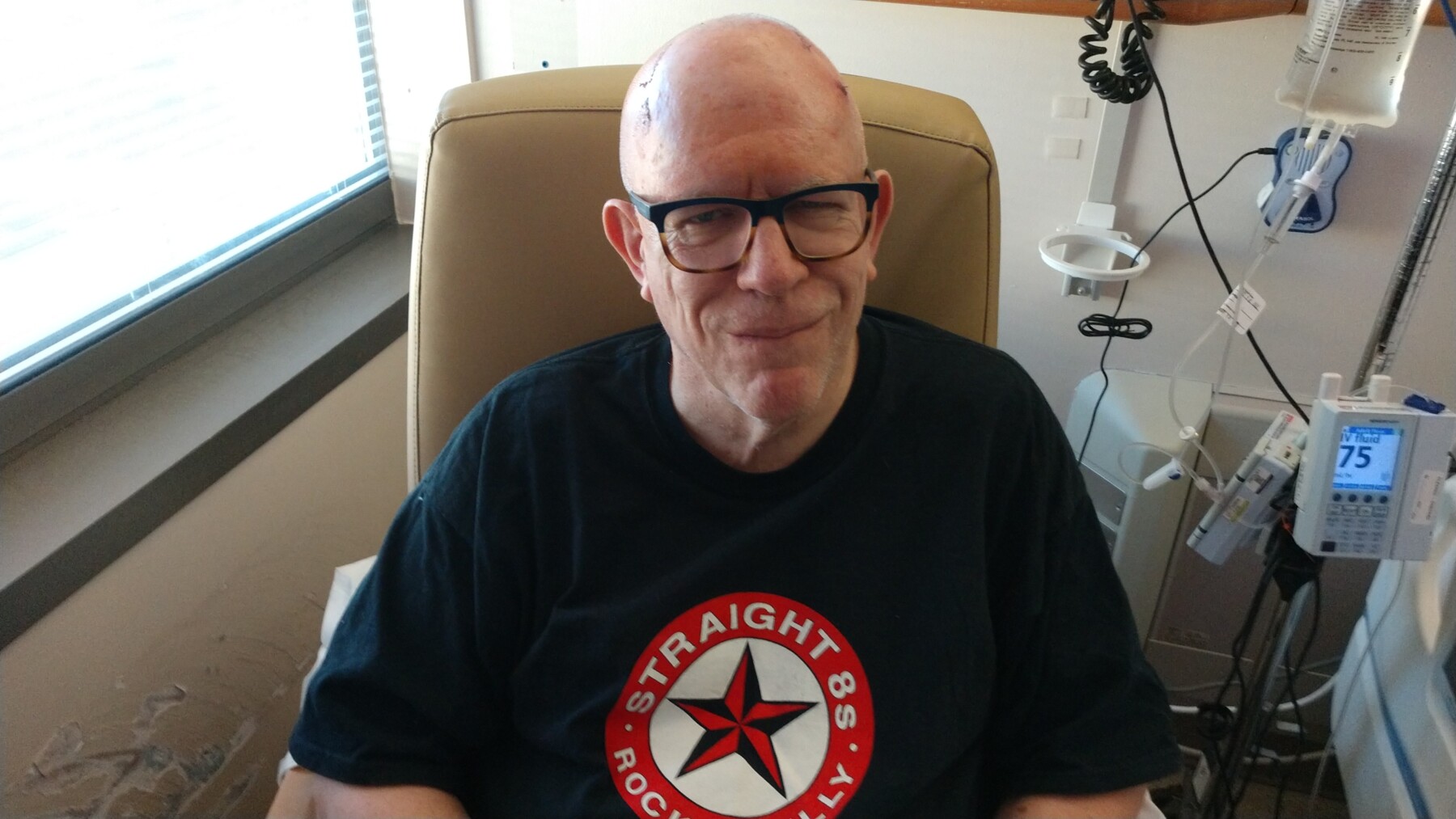 A man grins at the camera while seated in a chair in his hospital room. He's wearing glasses and a dark T-shirt, and we can see the ends of the incision on his head. There's a window to his right, and various pieces of medical equipment in the background.