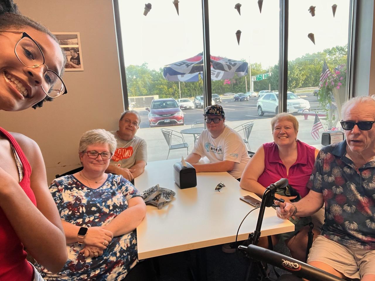 Six people are gathered around a table in what appears to be a restaurant. The columnist and her husband are on the right, with her husband seated behind his rollator.