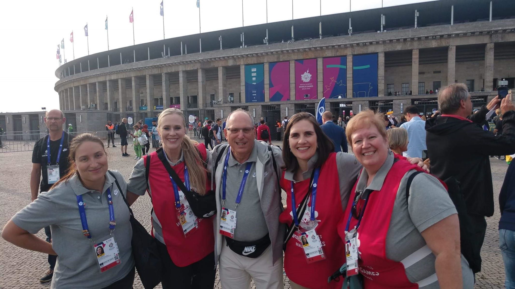 Five people — four women and one man — stand side by side with their arms around each other. They're all wearing gray collared shirts and blue lanyards, and three of the women have on red smocks. They're standing outside in front of some sort of large arena or coliseum. 