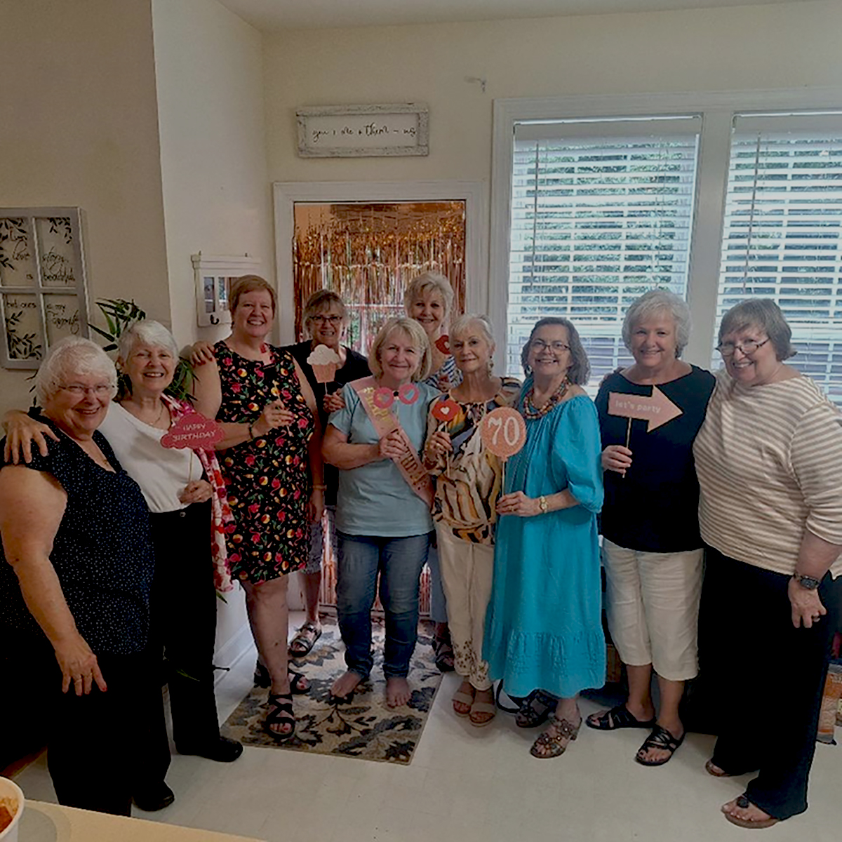 Ten older women stand in a U-shape in a living room or front room of an office or studio. One woman is holding a small sign with the number 70 on it, while another holds a pink arrow sign. It's not clear what these signs mean. 