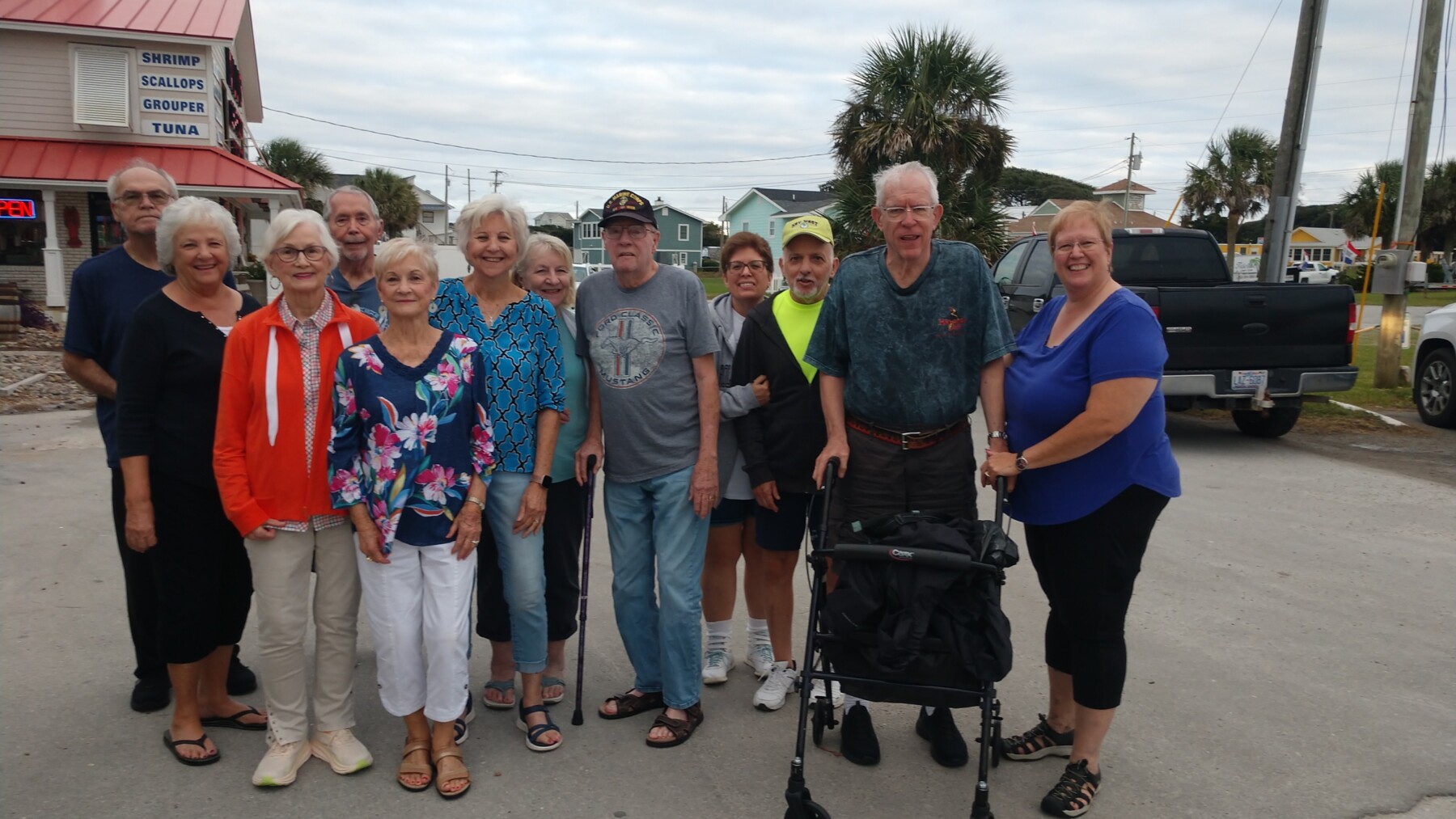 A group of 12 stand on a road beneath a cloudy blue sky. The man on the far right uses a mobility aid; another man has a cane.