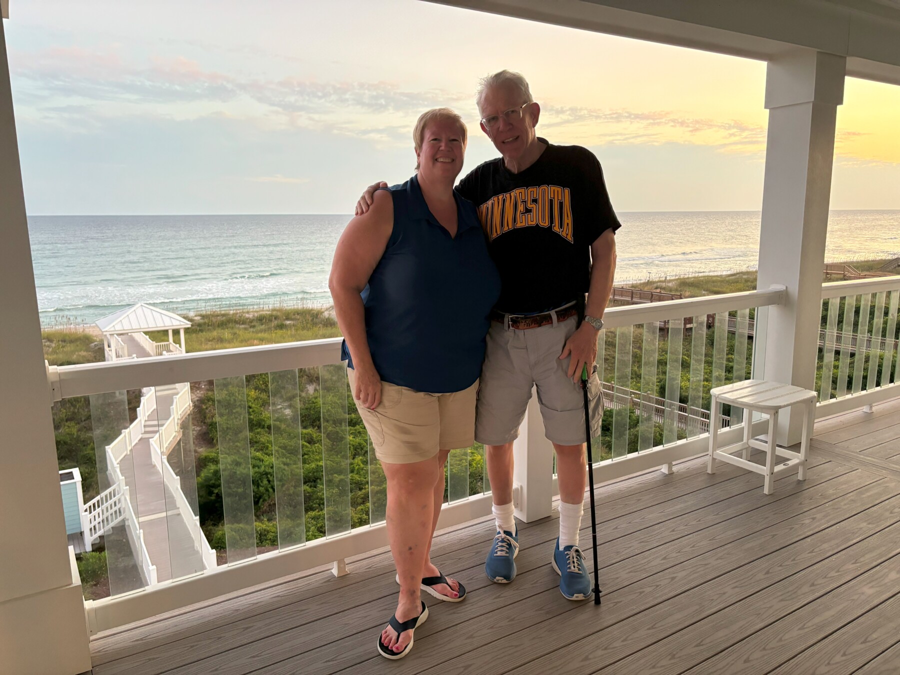 A married couple poses for a photo on a covered balcony that looks out over the beach. The woman is wearing a blue tank top and shorts, while the man is in a black "Minnesota" T-shirt and shorts. He has a walking stick in his left hand, and his right arm is draped around his wife's shoulders. The sun is setting over the ocean behind them.