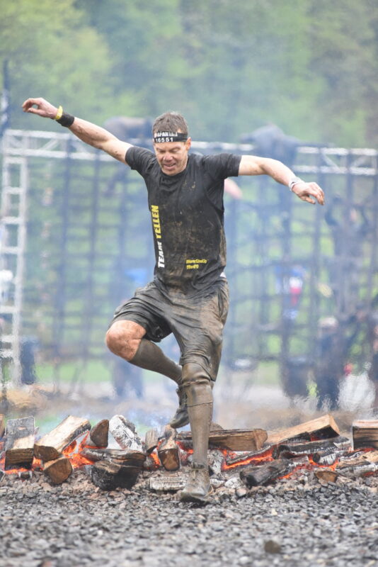 In a crazy photo, a muddied and sweaty man with a black headband and black T-shirt dramatically leaps over an obstacle course of burning firewood. 