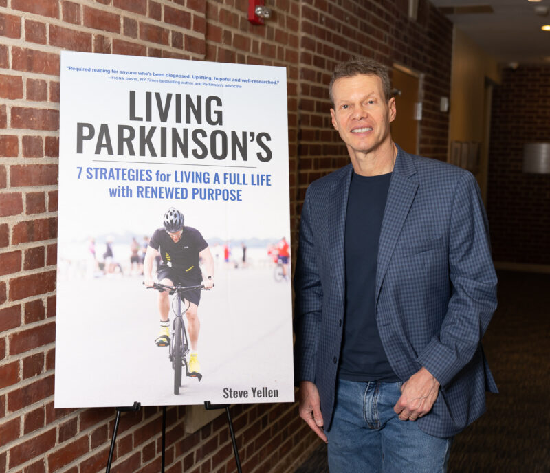 A proudly smiling middle-aged man in jeans and a checkered blue sport coat stands next to a poster of a book cover that shows a man on a bicycle and the book title: "Living Parkinson's: 7 Strategies for Living a Full Life with Renewed Purpose." 