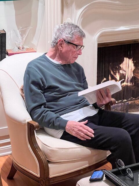 An older man sits in a reading chair by a fire in the fireplace, reading a book.