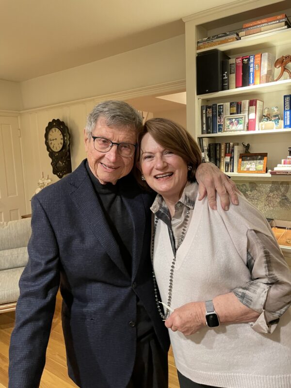 An older couple stands smiling in the living room of their home. Both are sharply dressed, and hugging each other. 